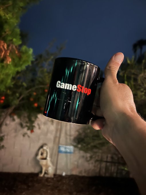 A hand holds up a black ceramic mug featuring the "GameStop" logo against a nighttime outdoor backdrop with trees and a statue.