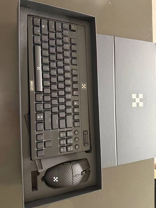 A black OKX-branded keyboard and mouse set sits in its branded packaging box featuring the OKX logo. The image shows the full-sized keyboard with its distinctive textured keycaps and a matching honeycomb-patterned gaming mouse, directly illustrating the peripheral gift mentioned in the post.