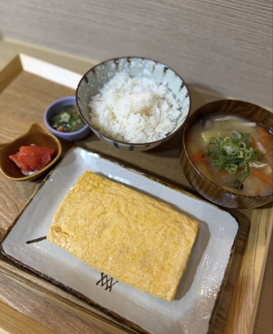 A traditional Japanese set meal featuring a thick tamagoyaki (rolled omelet) alongside white rice, miso soup, and small side dishes of pickled vegetables and a chili condiment, served on a wooden tray with plates bearing the logo "XXX."

The image illustrates the post's point about high Japanese restaurant prices, showing a relatively simple meal of eggs, rice, and soup that nonetheless costs 1,500 yen.