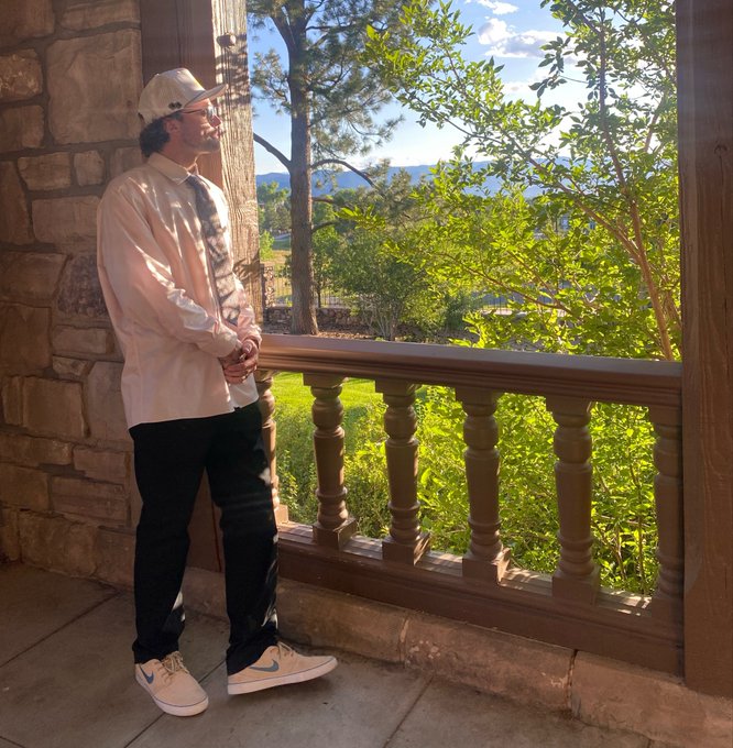A man in a beige cap, white button-up shirt, tie, and Nike sneakers leans against a stone balustrade, gazing out at a serene landscape of trees and distant mountains. The peaceful, reflective pose mirrors the "GM Web 3 I'm Longing $HYPE" post's vibe—evoking a quiet, longing moment amid bullish crypto enthusiasm.