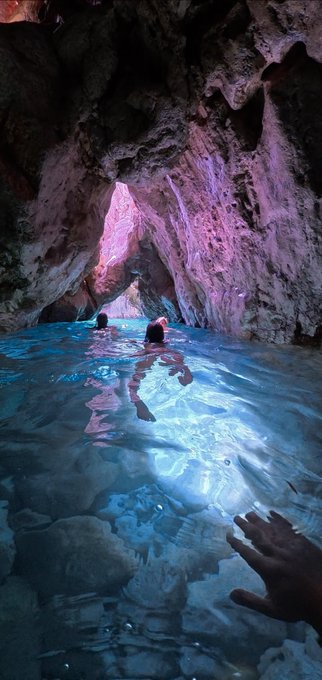 A moody photograph of people swimming inside a sea cave with glowing turquoise water and dramatic rocky walls, with one person's hand reaching out in the foreground.