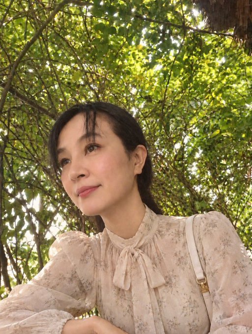A woman with dark hair poses outdoors in a floral blouse with a bow, with green leaves in the background.