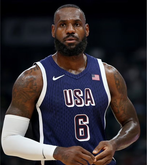 LeBron James wearing a blue USA basketball jersey with number 6, featuring an American flag patch and visible arm tattoos.