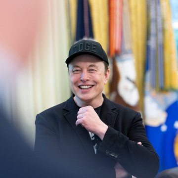 Elon Musk smiles broadly while wearing a black "DOGE" cap and dark jacket, with flags visible in the background. The image accompanies a quote in which Musk expresses cautious optimism about SpaceX developing a AI model comparable to GPT-6 within 2 to 3 months.