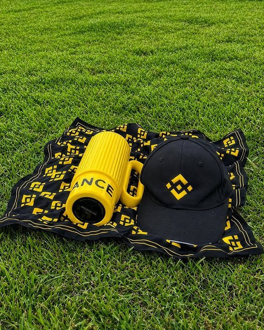 Un vaso amarillo de Binance, una gorra negra con el logo de Binance y una tela con motivos amarillos descansan sobre el césped. “Porque hasta el swag necesita un respiro” cobra sentido al mostrar la merchandising de Binance tomando un merecido descanso al aire libre.