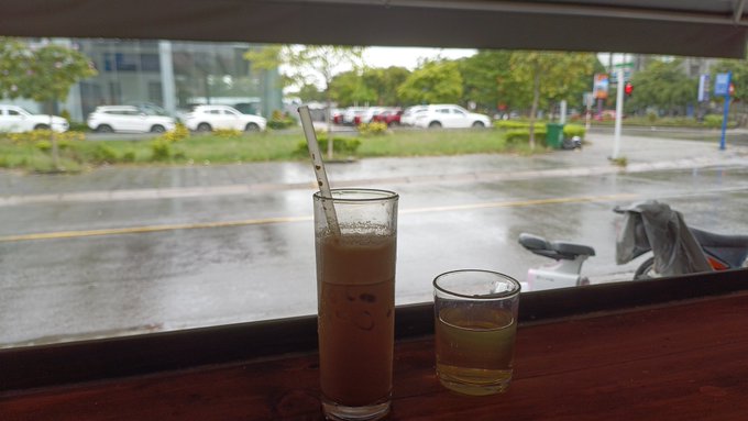 Two drinks—a tall glass of iced coffee with a straw and a smaller glass of what appears to be tea or a clear beverage—sit on a wooden windowsill overlooking a rain-soaked street with parked cars and a wet motorcycle in the distance, perfectly capturing the cozy rainy Sunday vibe of sipping coffee while scrolling through X and dreaming about landing a Web3 job.