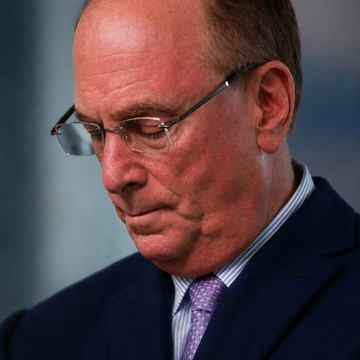 A close-up portrait of an older man with glasses, wearing a dark suit and a purple tie, looking downward.