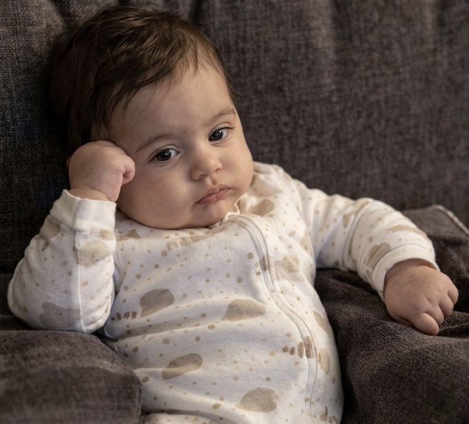 Close-up photograph of a baby with dark hair wearing a cream-colored onesie with beige spots, lying against a brown textured fabric.