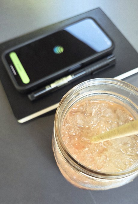 A glass jar filled with a light-colored beverage and ice sits next to a black notebook, a pen, and a smartphone with a green case displaying a logo.