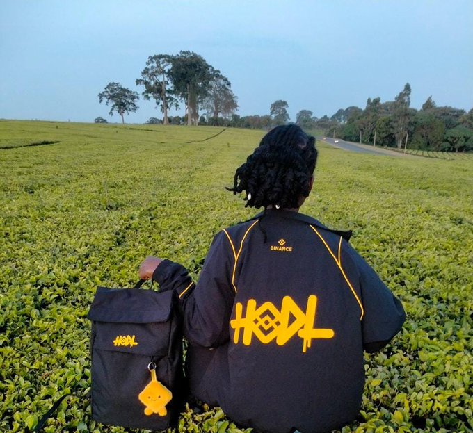 A person stands in a green tea field wearing a black jacket with yellow 'HODL' graffiti-style lettering and a 'BINANCE' logo, holding a black backpack with a matching yellow character charm.