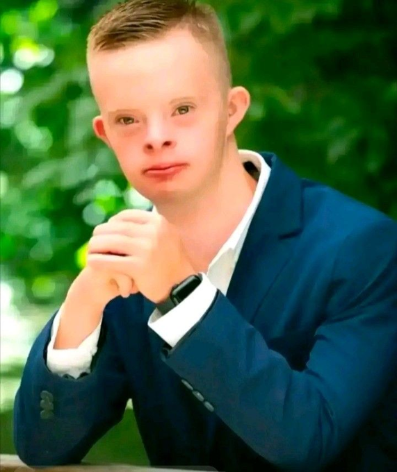 A portrait of a young boy with short blonde hair wearing a blue suit jacket and white shirt, sitting with his hands clasped together outdoors.