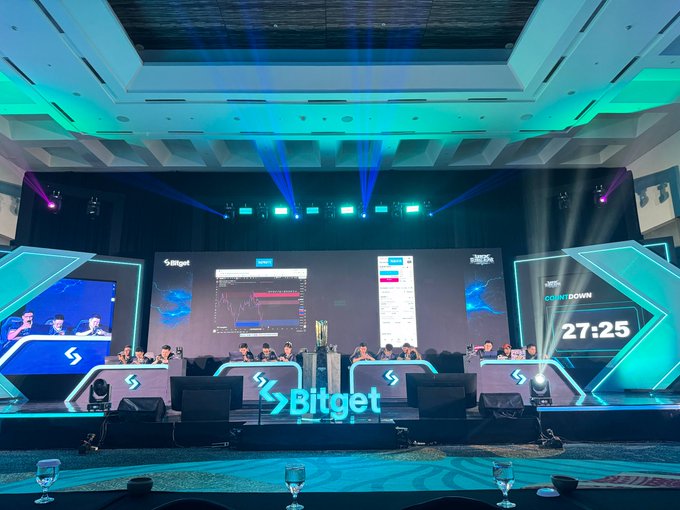 The image shows a large e-sports style trading competition stage for Bitget's UEX GAT (Global Account Trading) competition, featuring illuminated "Bitget" branding and a countdown timer displaying "27:25" alongside multiple traders competing at their stations. The dramatic blue and cyan lighting, overhead spotlights, and arena-like setup support the post's description of it feeling like an international "battle arena" rather than a typical trading contest.