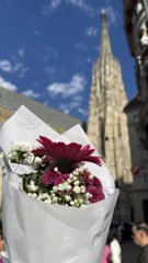 A bouquet of red and white flowers wrapped in white paper is held up against a backdrop of a Gothic-style cathedral spire under a partly cloudy sky.