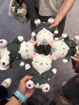 A photo of a circular arrangement of white cat plush toys dressed in green military-style uniforms, with hands arranging them in a circle on a floor.