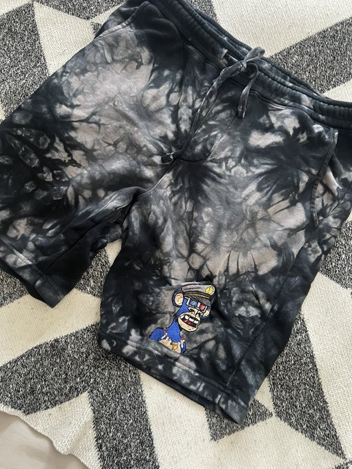 A pair of black tie-dye shorts featuring a Bored Ape Yacht Club character patch rests on a checkered rug, showcasing the custom DIY design referenced in the caption. The embroidered blue ape graphic at the hem represents the creator's personalized NFT apparel project.