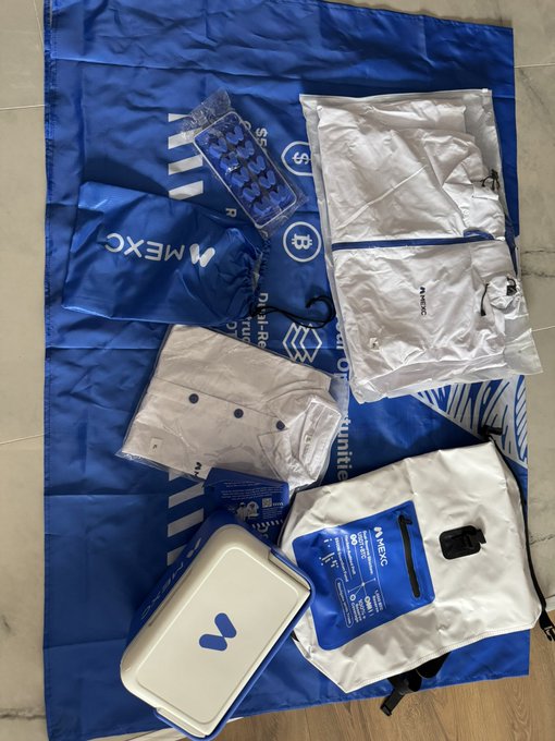 A flat lay of Mexc-branded merchandise and giveaways on a tiled floor, including a blue Mexc drawstring bag, a Binance-themed towel/poster, white shorts, a pill blister pack, and other promotional items.