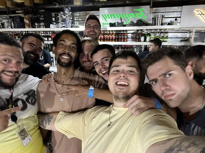 A group of young men pose together for a selfie inside what appears to be a bar or restaurant with shelves of bottles in the background.