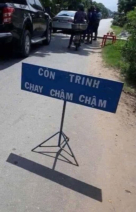 A blue road sign on a metal stand reads 'CÒN TRÌNH CHẠY CHẬM CHẬM' in Vietnamese, placed on a road with vehicles and pedestrians in the background.