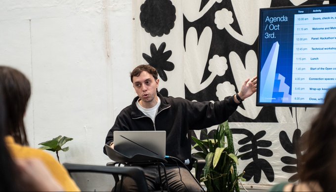 A man sitting with a laptop in front of a screen displaying 'Agenda / Oct 3rd' with a schedule, gesturing while speaking to an audience in a room with a black and white floral wall hanging.