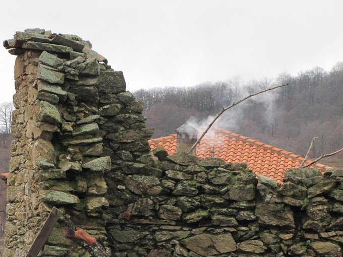 A weathered stone wall in the foreground with a terracotta-tiled roof and a smoking chimney behind it, set against a backdrop of bare winter trees under a cloudy sky.
