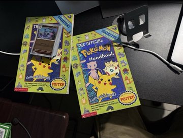 Two copies of 'The Official Pokemon Handbook' by Totter Books sit on a desk, featuring a Pokedex in the box packaging.