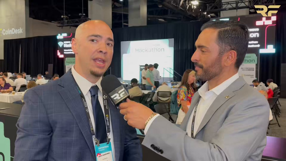 The image shows two men dressed in suits talking into a microphone during an event or gathering. One man appears to be wearing a name tag that reads "@RayFuentesIO" while the other has a name tag that says "@consensus2026". They are discussing various topics, including XRP and institutional adoption of cryptocurrencies like Bitcoin. The men seem to be engaged in a conversation about the future direction of the crypto market.