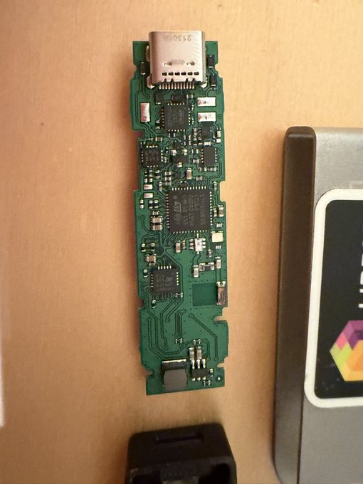 A green circuit board from a disassembled electronic device, featuring a USB-C port at the top and various microchips, sitting on a wooden surface next to another component.