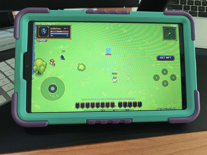 An iPad in a teal and purple protective case displays a video game featuring a character named "CableUser" on a green landscape, with a "GET NFT" button visible on screen.