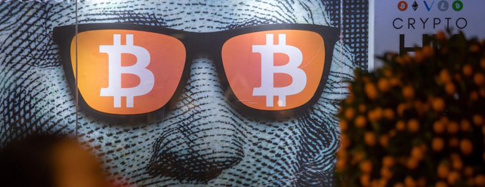 A mural or graphic shows a face wearing sunglasses with the Bitcoin 'B' logo reflected in each lens, with a 'CRYPTO' sign partially visible.