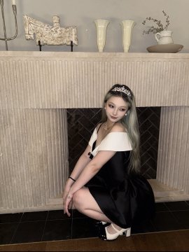 A photo of a young woman wearing a tiara and an elegant off-shoulder black and white dress, posing in front of a white marble fireplace.