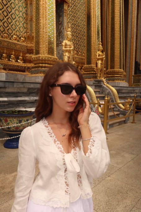 A young woman with long brown hair wearing sunglasses and a white lace top poses in front of an ornate golden temple structure with intricate decorative details.