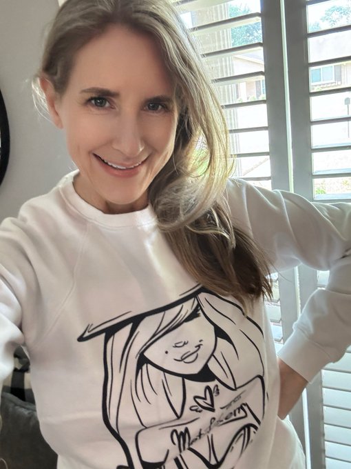 A selfie of a smiling woman with long blonde hair wearing a white sweatshirt featuring a black line drawing of a girl.