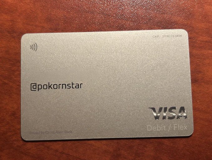 A sleek metallic Visa Debit/Flex card with "@pokornstar" embossed on it rests on a wooden surface, representing the user's new modern checking account with X Money that they signed up for after not having a new account since 2008. The minimalist card design reflects the "stepping into the future" theme of upgrading from an older banking setup.