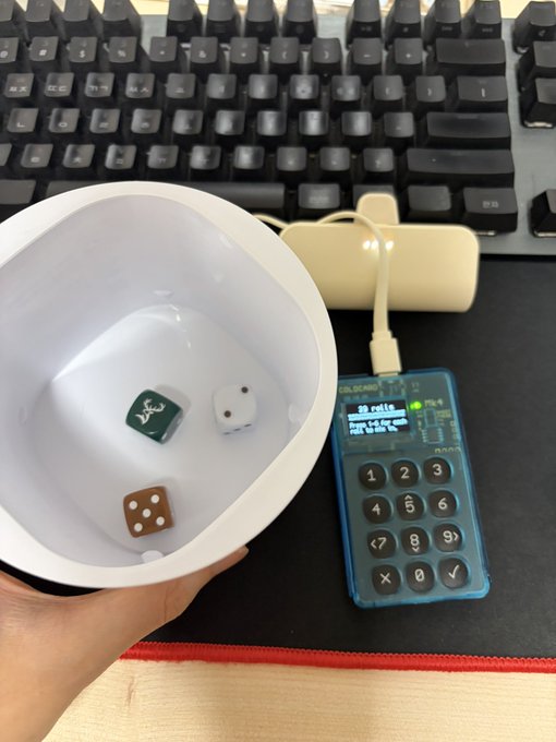 A hand holds a white bowl containing three dice next to a computer keyboard and a blue electronic calculator-style device displaying "20 rolls".