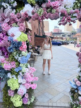The image features a woman standing in front of an archway made up entirely of flowers, with her back facing the camera. The archway is adorned with various types of colorful flowers, creating a beautiful and vibrant scene. In the background, there are several cars parked along the street, indicating that it might be a busy area or a popular location for visitors to enjoy the floral display.