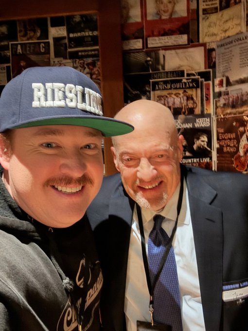 A selfie of two smiling men, one wearing a 'RIESLING' cap, standing in front of a wall covered in country music posters including 'BRANSCO' and 'EMPIRE BLUES'.