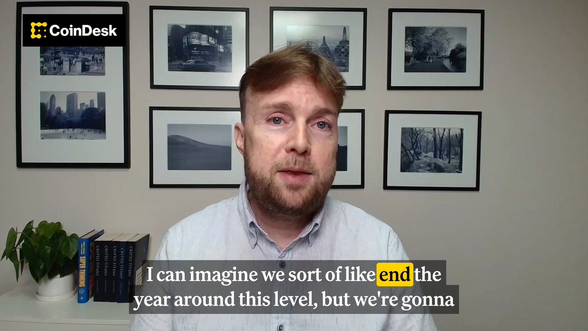 A bearded man in a light blue shirt speaks in front of a wall of framed photos, with a "CoinDesk" logo in the corner and a book set titled "Crypto Titans" visible on a shelf. Subtitles read "I can imagine we sort of like end the year around this level, but we're gonna," aligning with the post's prediction of Bitcoin potentially ending the year around $60,000-$65,000 after a dip.