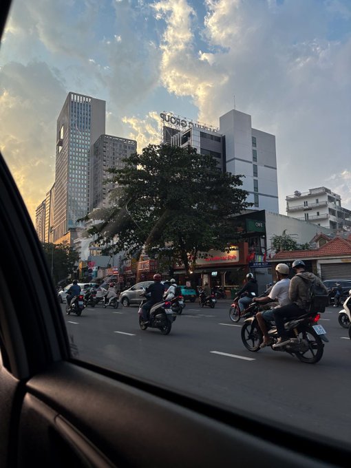 A view from inside a car of a busy city street in Vietnam, showing motorcyclists, the 'ĐẢNG CỘNG SẢN' building, and a sunset sky.
