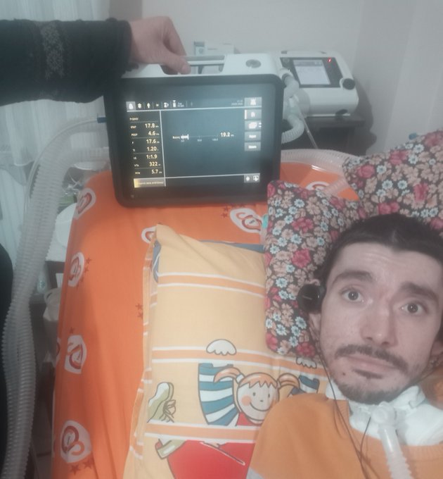 A man lies in a hospital bed with breathing tubes, while a hand holds up a medical monitor displaying vital signs.