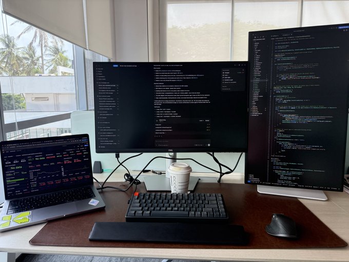 A developer's workstation featuring a laptop and two vertical monitors displaying terminal and code editor windows, accompanied by a mechanical keyboard, ergonomic mouse, and a coffee cup on a desk mat.
