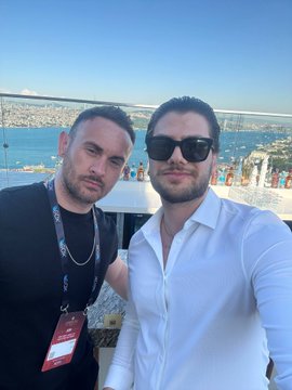 The image features two men posing for a photo, both wearing sunglasses and standing next to each other against a backdrop of water and a city skyline. One man has a red lanyard around his neck, while the other wears a white shirt. The text "Solana Summer" is visible in the image, which could be related to the location or event they are attending.