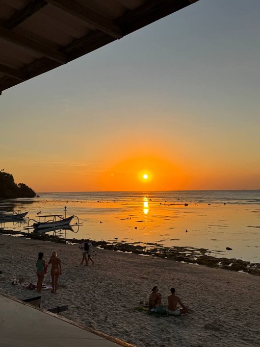 A calm Balinese beach at sunset, with people relaxing on the shore, boats in the shallows, and the sun reflecting across the water. The view captures the peaceful local atmosphere described after arriving for Coinfest Asia on August 20–21.