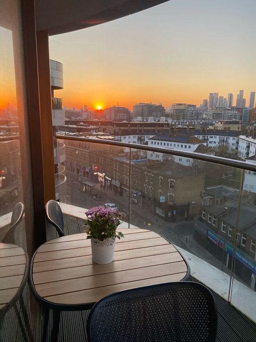 A sunset view from a balcony featuring a small wooden table with a potted pink flower, overlooking a cityscape with the Canary Wharf skyline visible in the distance.
