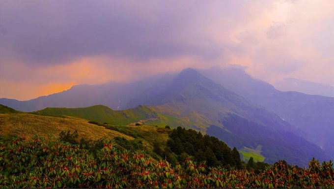 A mountain landscape at sunset, with dramatic purple and orange clouds hovering over rolling green peaks and shrubs in the foreground. The serene, uplifting scene complements the message of resilience and trust, symbolizing the calm and beauty that comes after persevering through challenges.