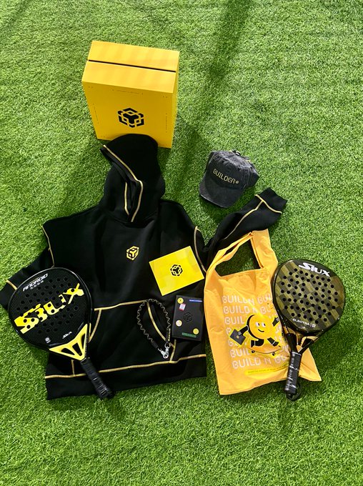A flat lay on artificial green grass showing paddle/padel equipment: two Siux padel rackets, a yellow Builder Box, a black and yellow hoodie and cap branded 'BUILDER', a yellow tote bag reading 'BUILD IT OWN IT', and tags/cards.