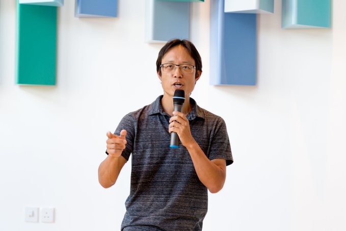A man with glasses and a dark polo shirt speaks into a microphone while gesturing, standing before a wall decorated with teal and blue rectangular panels. The image relates to the post as it likely depicts Yat Siu, the crypto executive mentioned, delivering a talk on rekindling excitement in the crypto space through NFTs, the metaverse, and gaming.