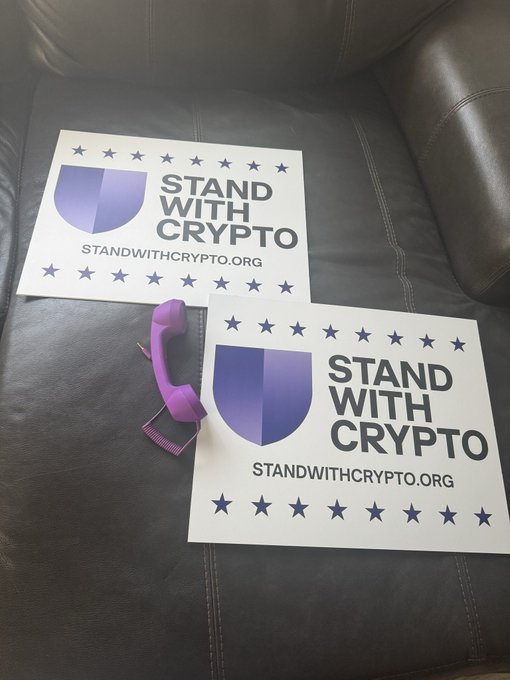 Two 'Stand With Crypto' stickers from standwithcrypto.org placed on a dark leather couch next to a purple hair clipper.