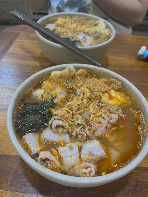 Two bowls of noodle soup topped with meat, fried shallots, egg, and greens sit on a wooden table with chopsticks resting on the rim of the back bowl.