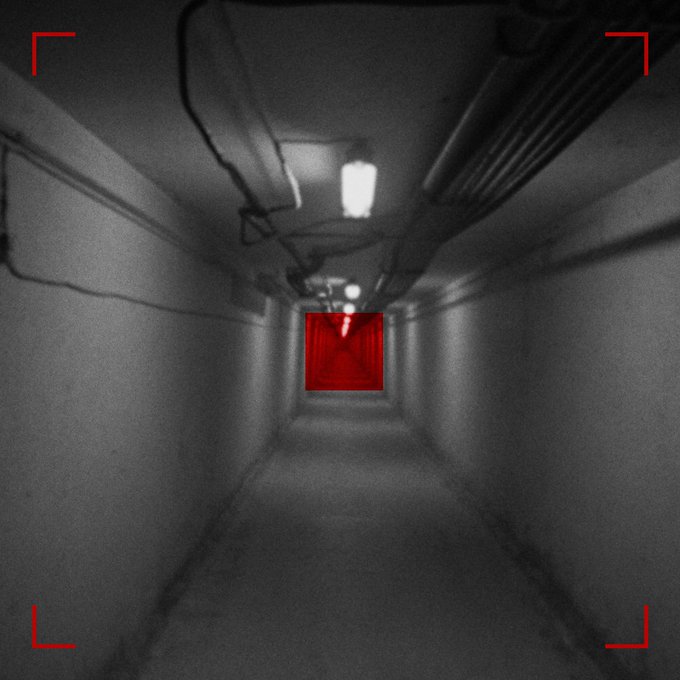 A dark, grainy surveillance-style view down a narrow hallway with pipes running along the ceiling, a bright light in the distance, and a red-lit doorway at the end, framed by red corner brackets.