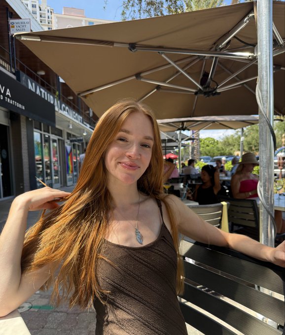 A young woman with long reddish-brown hair sitting at an outdoor cafe under a beige umbrella, wearing a dark top and a feather necklace, with storefronts visible in the background.